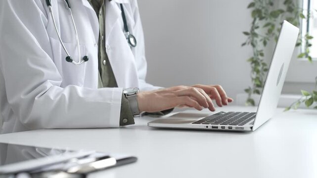 Medical professional wearing white lab coat typing on laptop, documenting patient information with clinical precision at workstation. Medicine and health care concept