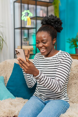 African American woman celebrating good news on smartphone. Sitting at home sofa, joyful...