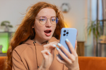 Surprised Caucasian redhead woman with windswept hair using smartphone, receives mail good news...