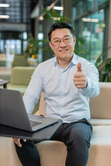 Naklejka premium Like. Happy young businessman guy working on laptop looking approvingly at camera showing thumbs up, like sign positive something good, positive feedback sitting in modern office lobby. Businesspeople