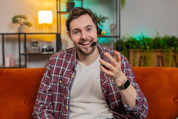 Call me. Happy Caucasian young man sitting on sofa at home wearing headset freelance worker call...