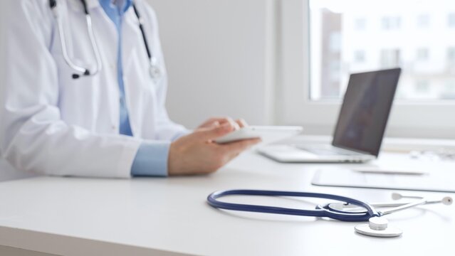 Closeup of blue stethoscope is lying on the table near female doctor reviewing patient data on tablet, representing digital healthcare workflow in contemporary medical setting