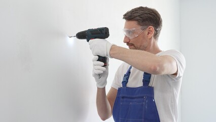 Middle aged man wearing blue overall, is drilling hole in white wall, wearing protective gear...