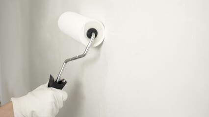 Close up focusing on gloved hand rolling white paint across wall surface, applying smooth, even...