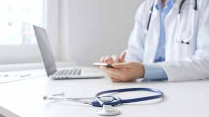 Closeup of blue stethoscope is lying on the table near female doctor checking smartphone...