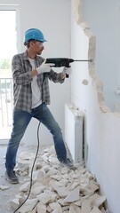 Male construction worker wearing grey checked shirt and blue hardhat is demolishing a wall with a...