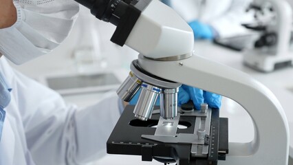 Close up of scientific professional blue gloved hands examining microscope lens, performing...