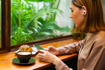 Beautiful Asian woman sitting by the window in a cozy café, enjoying coffee and pastry, listening...