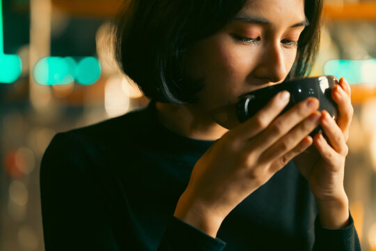 Young Asian woman sitting counter bar drinking a cup of coffee at cafe coffee shop in dark lighting. Portrait of Attractive girl enjoy indoor lifestyle cozy relaxing casual urban stylish with coffee.