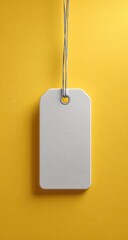 A blank, off-white rectangular tag hangs from a thin, grey string against a vibrant yellow backdrop.  The tag features a small, metallic eyeleted hole at the top