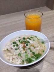 Minced chicken congee topped with chopped spring onions and fried garlic, served with orange juice as breakfast on wooden table, Bangkok, Thailand 