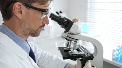 Medical scientist wearing gloves and protective glasses examining samples under a microscope in a...
