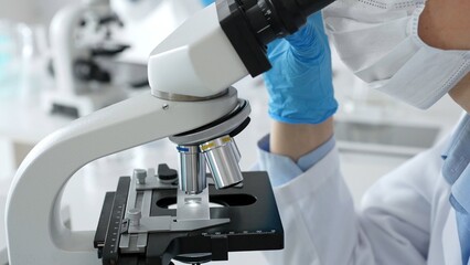 Close up of scientific professional blue gloved hands examining microscope lens, performing...