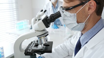 Masked scientist analyzing sample under microscope, colleague researching nearby in sterile medical...