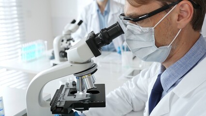 Masked scientist analyzing sample under microscope, colleague researching nearby in sterile medical...