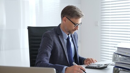 Mature entrepreneur in suit, tie and glasses is sitting at desk, writing notes and using calculator...