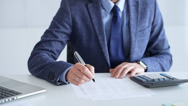 Businessman wearing suit and tie is using calculator and taking notes while working at desk in office, with laptop and stack of folders nearby, close up. Audit and taxes theme in business