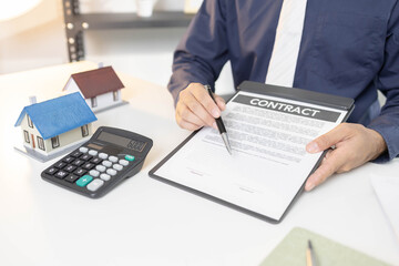 A man is sitting at a desk with a laptop and a notebook. He is writing in the notebook and has a pen in his hand. There are three houses on the desk, and a calculator is also present
