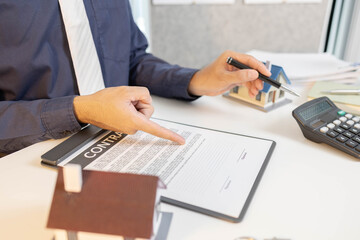 A man is sitting at a desk with a laptop and a notebook. He is writing in the notebook and has a pen in his hand. There are three houses on the desk, and a calculator is also present
