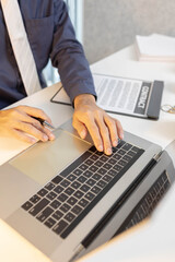 A man is sitting at a desk with a laptop and a notebook. He is writing in the notebook and has a pen in his hand. There are three houses on the desk, and a calculator is also present
