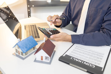 A man is sitting at a desk with a laptop and a notebook. He is writing in the notebook and has a pen in his hand. There are three houses on the desk, and a calculator is also present

