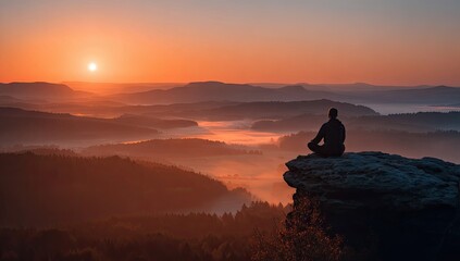 Obraz premium Silhouetted man meditates atop a rocky outcrop at sunrise, overlooking a misty valley and distant mountains bathed in warm, orange light