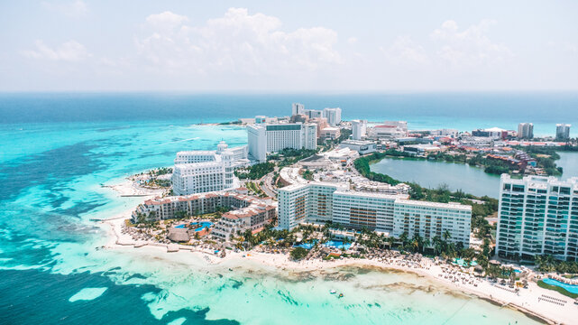 A beautiful beach with a city in the background. The city is full of tall buildings and the beach is full of people. The sky is clear. Aerial Views of Cancun Hotel zone, Quintana Roo Mexico 