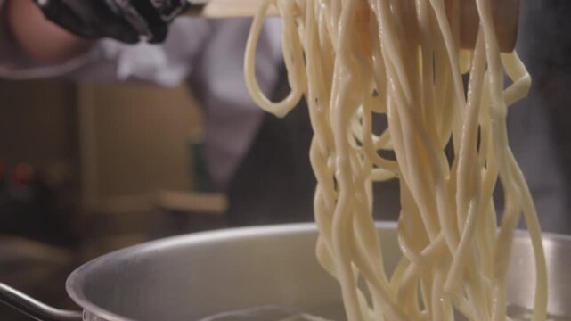 Close-up hands of chef in gloves and apron while stirring handmade pasta in the saucepan with boiling water. Restaurant of traditional Asian kitchen.
