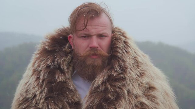 A manly man thick red beard wearing a fur coat looks at the camera standing in the snow. Male face with serious expression. Viking character, strength and protection.