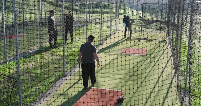 White male batter swinging bat in batting cage as pitcher winding arm, hurling ball for practice