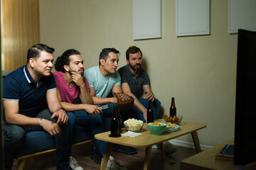 Latin male friends watching sports on tv at home, drinking beer