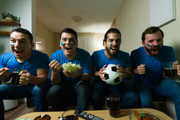 Excited male friends cheering and drinking beer watching soccer match at home