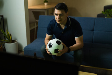 Latin man holding soccer ball watching game on tv at home