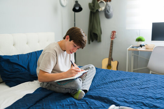 Male teen writing on notebook sitting on bed in his bedroom