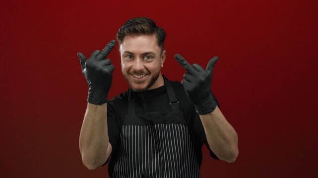 Young man with beard wearing gloves and apron over red background making inappropriate hand gestures with a defiant expression, showcasing boldness and attitude.