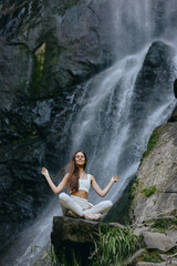 young woman meditating near waterfall in nature, peaceful, calm, yoga practice by rocks and flowing...