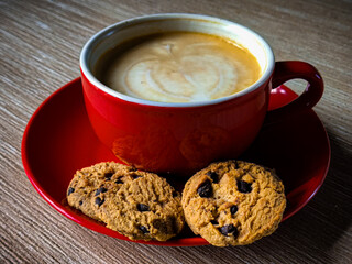 An overhead photo showing a cup of coffee with latte art on a red plate, accompanied by two chocolate cakes on a wooden table.