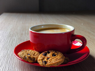 A close-up shot shows a cup of hot coffee with a bit of latte art on top, accompanied by a chocolate chip cookie snack. All objects are placed on a wooden table, creating a warm and cozy atmosphere.