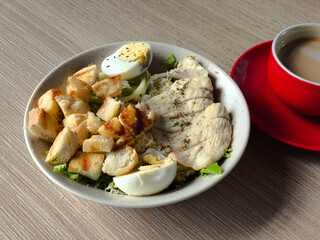 A photo of a Caesar salad served in a bowl on a wooden table, with a cup of coffee on the right. The dish includes sliced chicken breast, a hard-boiled egg, and toast (croutons).