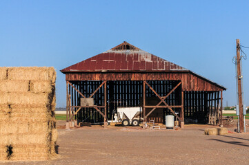 A wooden open-air barn with a rusted metal roof is used for storing hay bales and farm equipment, representing traditional agricultural infrastructure on a rural farming property