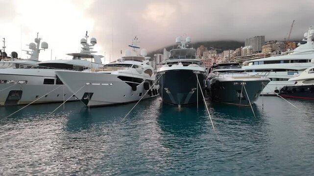 Row of sleek luxury yachts, docked in Port Hercules with the Monaco cityscape and misty hills in the background