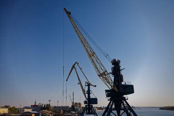 Industrial crane towering over docks in vibrant blue sky setting. A towering gantry crane commands the foreground of this bustling waterfront, extending over calm waters. Behind it, shipping container