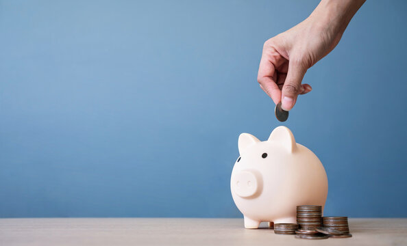 Hand of woman putting coin into piggy bank on a wooden table with blue background. saving money and invest concept.
