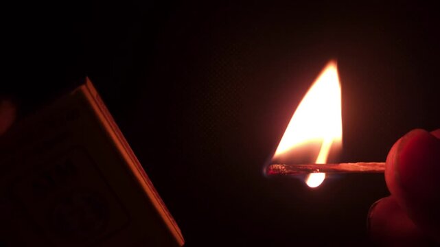 Extreme close-up captures fingers lighting a match, the sulfur tip igniting with fire and smoke, burning brightly in the darkness.