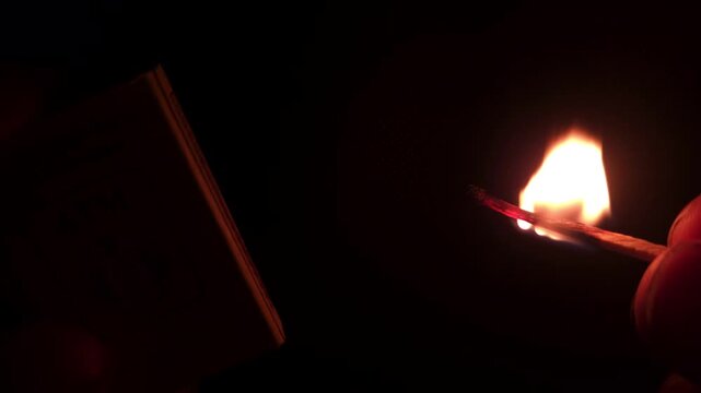 Extreme close-up captures fingers lighting a match, the sulfur tip igniting with fire and smoke, burning brightly in the darkness.
