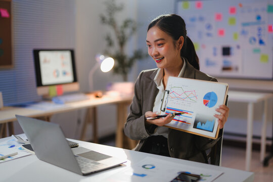 Young professional explaining diagrams and graphs on a clipboard during a video call in her office at night - Powered by Adobe