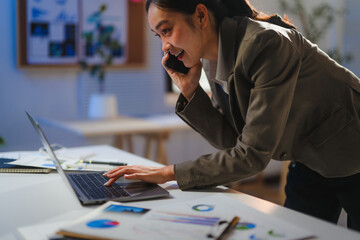 Young manager working late at night, using laptop and talking on smartphone, analyzing financial charts and graphs
