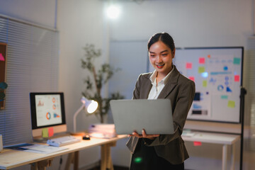 Young manager standing and using laptop, working late at night in office with charts and graphs in background