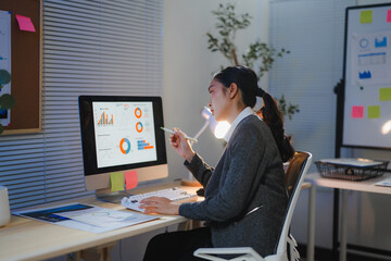 Businesswoman analyzing financial charts and data on computer, working late in modern office using computer and pen