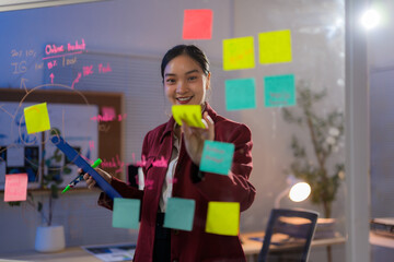 Asian businesswoman attaching sticky notes on glass wall and writing, working and planning strategy in office at night